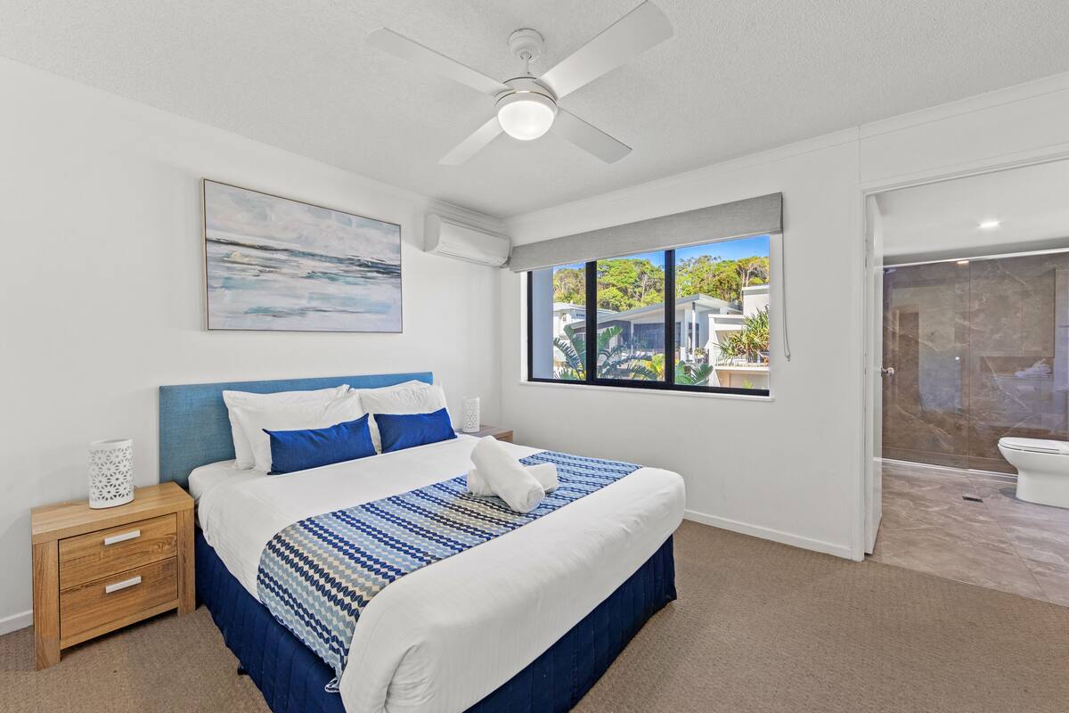 A bedroom with a bed, a nightstand, a ceiling fan, a window with a view, and a bathroom with a toilet.