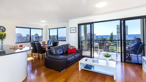 A modern living room with a black leather sofa, a white coffee table, and a dining area with a white table and chairs. Large windows offer a view of the city and ocean. - The Mirage Alexandra Headland (Alexandra Headland)
