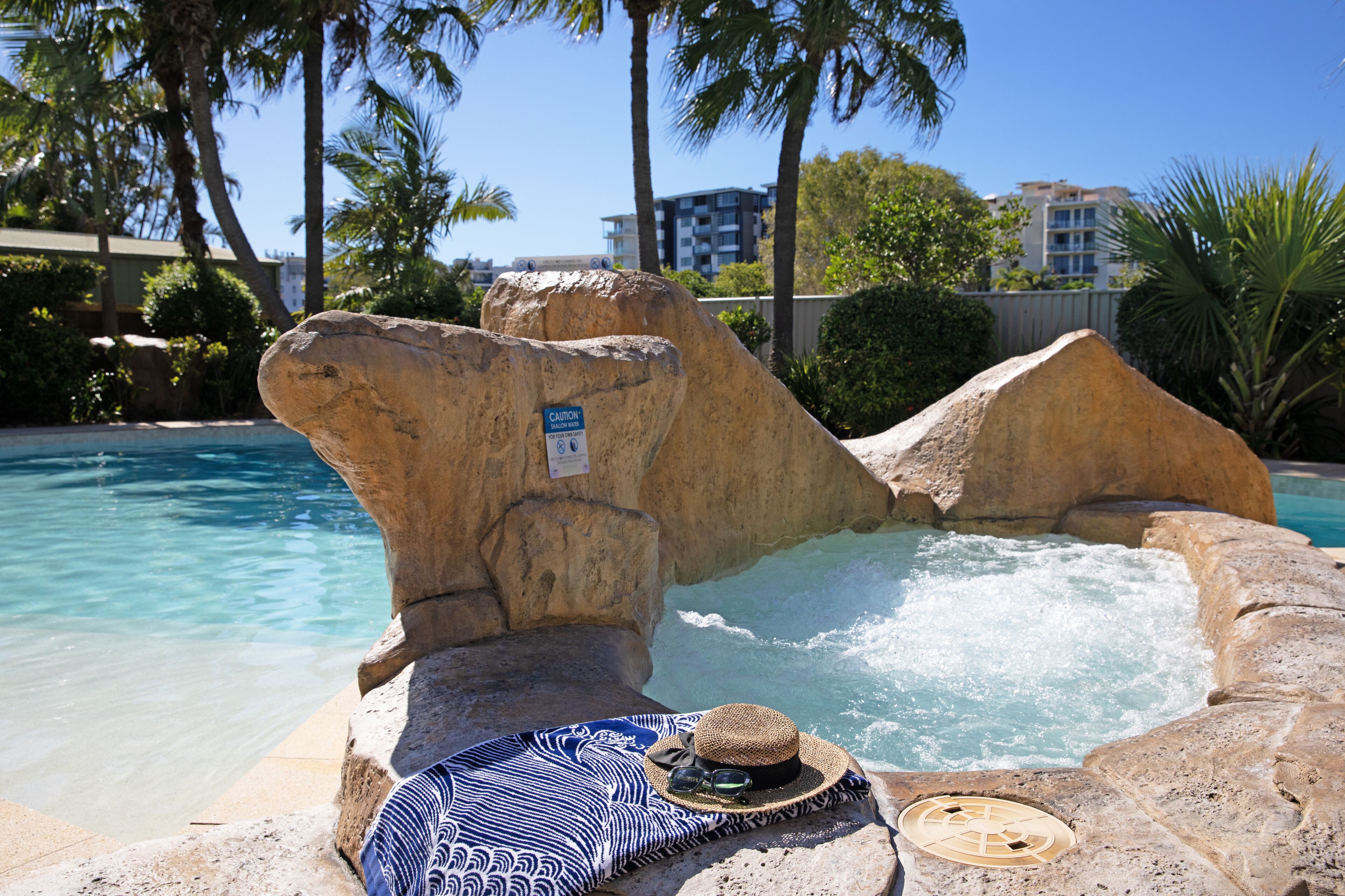 A hot tub surrounded by large rocks and palm trees, with a pool in the background.