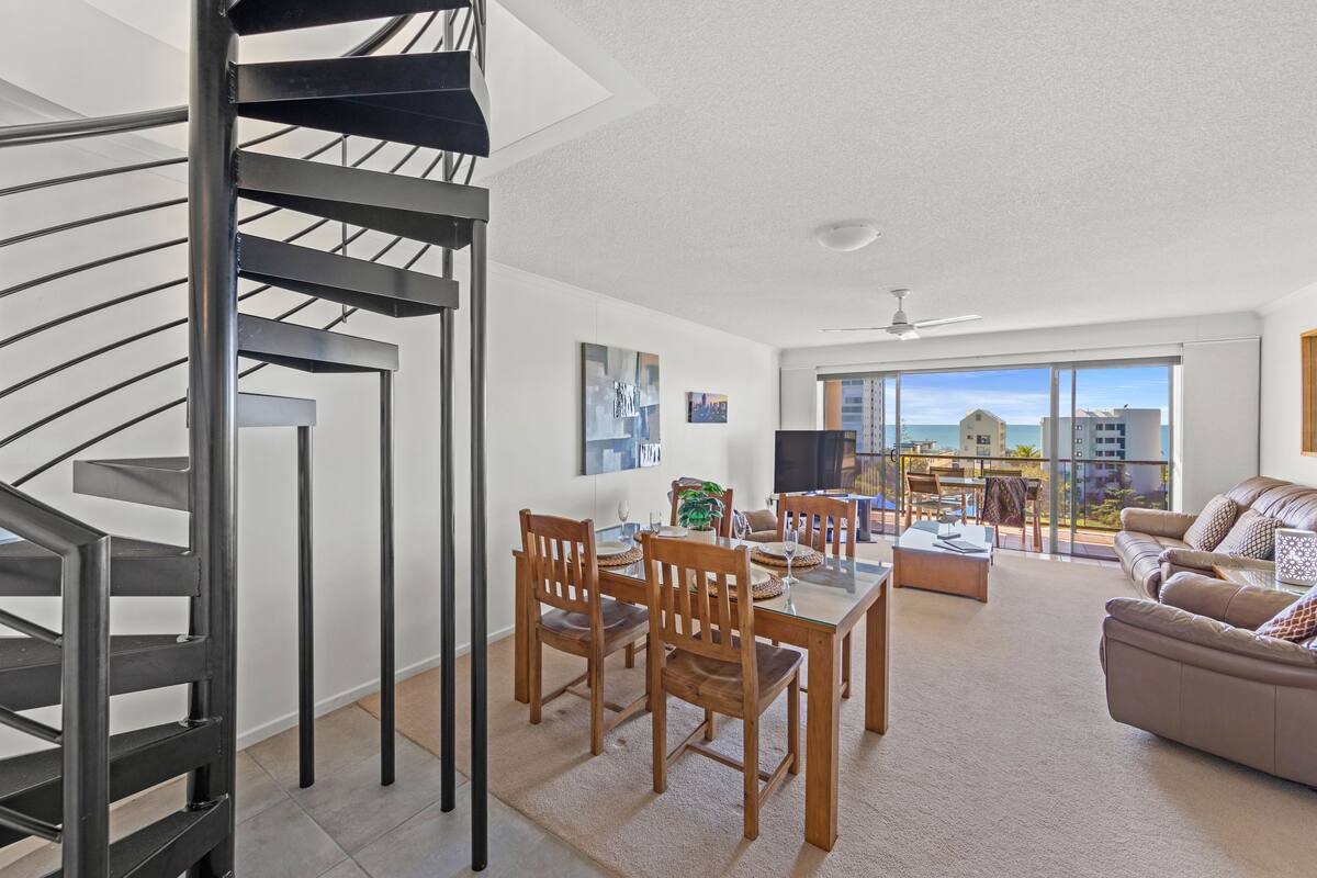 A modern living room with a dining area, a spiral staircase, and a balcony with a view of the sea.