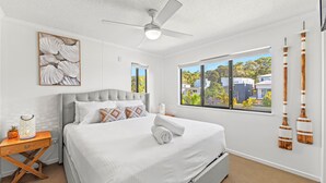 A modern bedroom with a large bed, a ceiling fan, a window with a view of buildings, and decorative wall art. - The Mirage Alexandra Headland (Alexandra Headland)