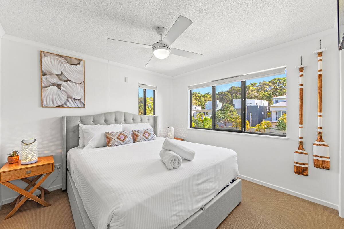 A modern bedroom with a large bed, a ceiling fan, a window with a view of buildings, and decorative wall art.