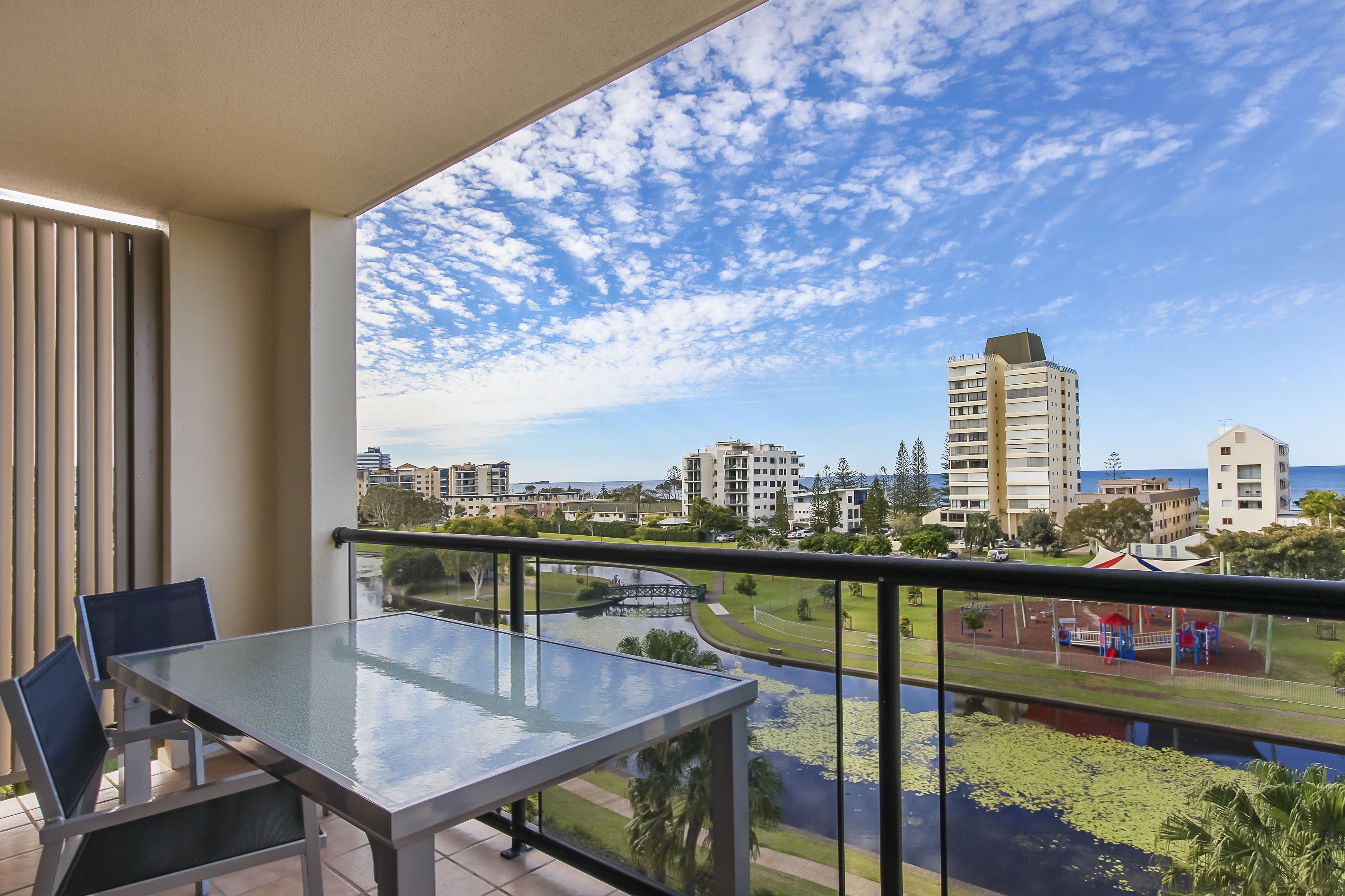 Een balkon met een glazen tafel en stoelen, met uitzicht op een stadsbeeld met gebouwen en een park.