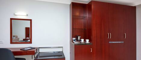 A room with a wooden wardrobe, a desk with a chair, a bench, a mirror, and a small shelf with a kettle and cups.