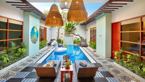 A swimming pool with a swan float, surrounded by a wooden deck and wicker chairs, with hanging rattan lights and a tropical garden. - The Vie Villa Legian (Seminyak)