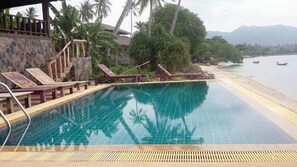 A swimming pool with lounge chairs, palm trees, and a view of the beach and mountains.