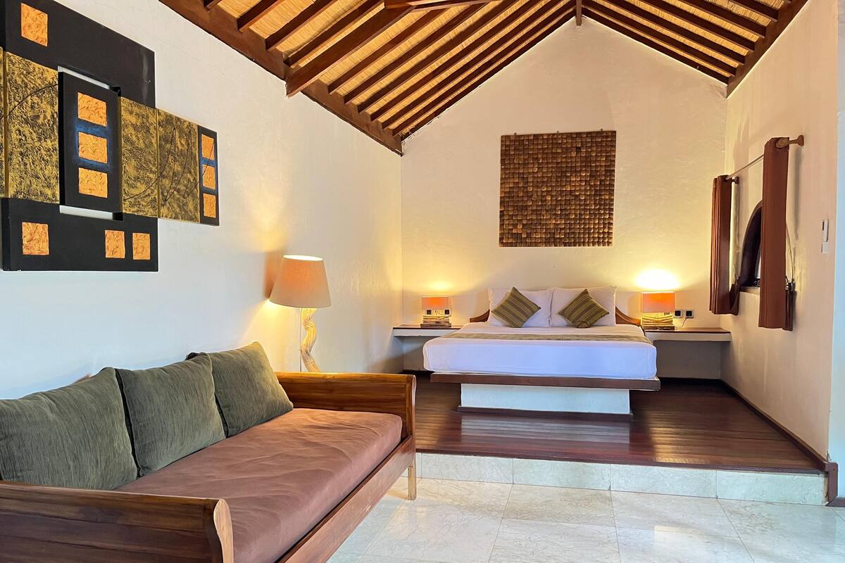 Een hotelkamer met een houten plafond, een bed met wit beddengoed, een houten bankje en een wandlamp.