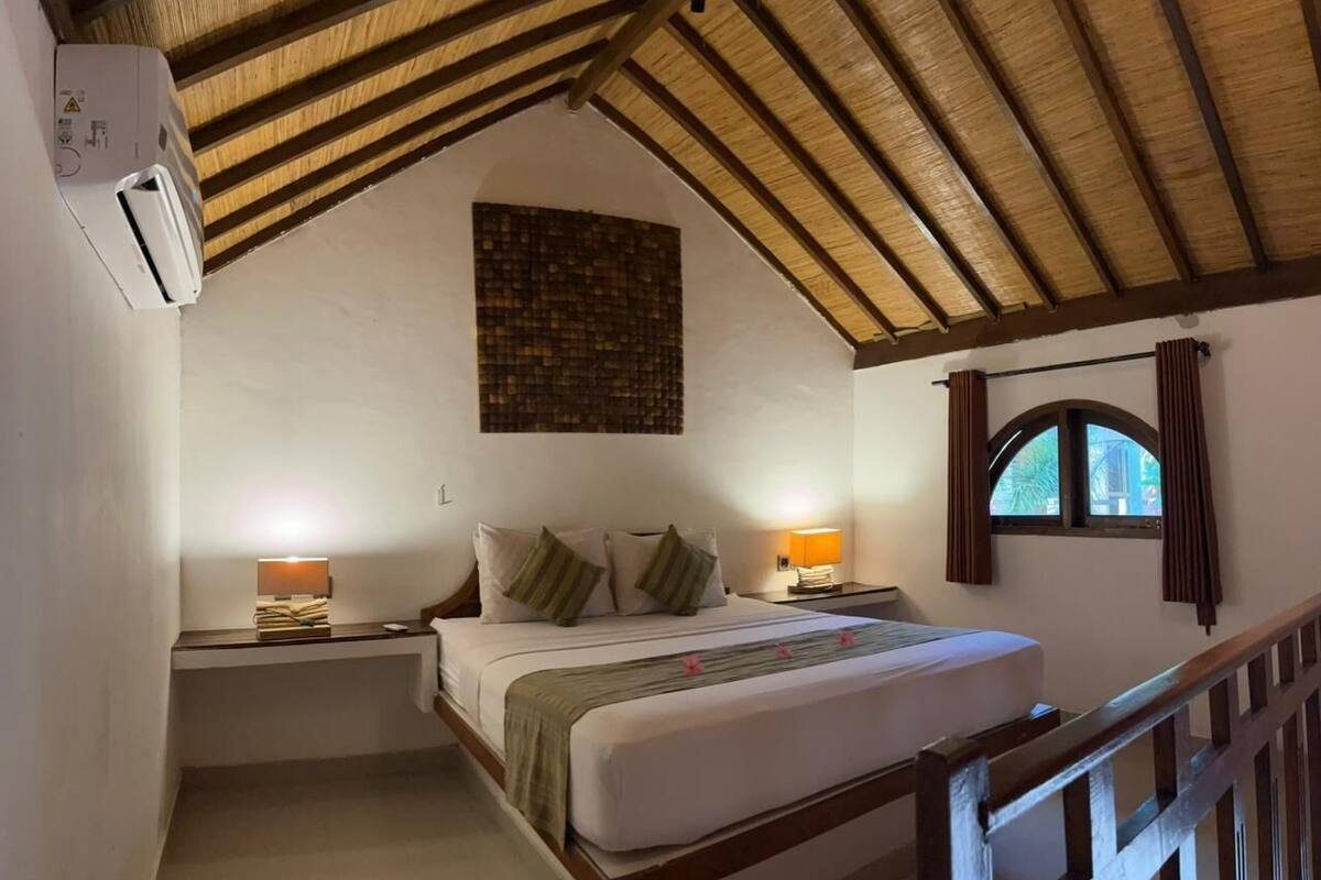 Een slaapkamer met een schuine kap, een bed met kussens, nachtlampjes en een raam met gordijnen.