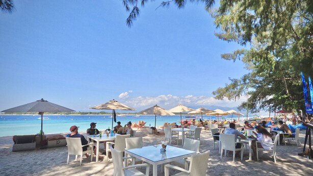 A beachside café with outdoor seating, tables, and umbrellas, overlooking a clear blue sea.