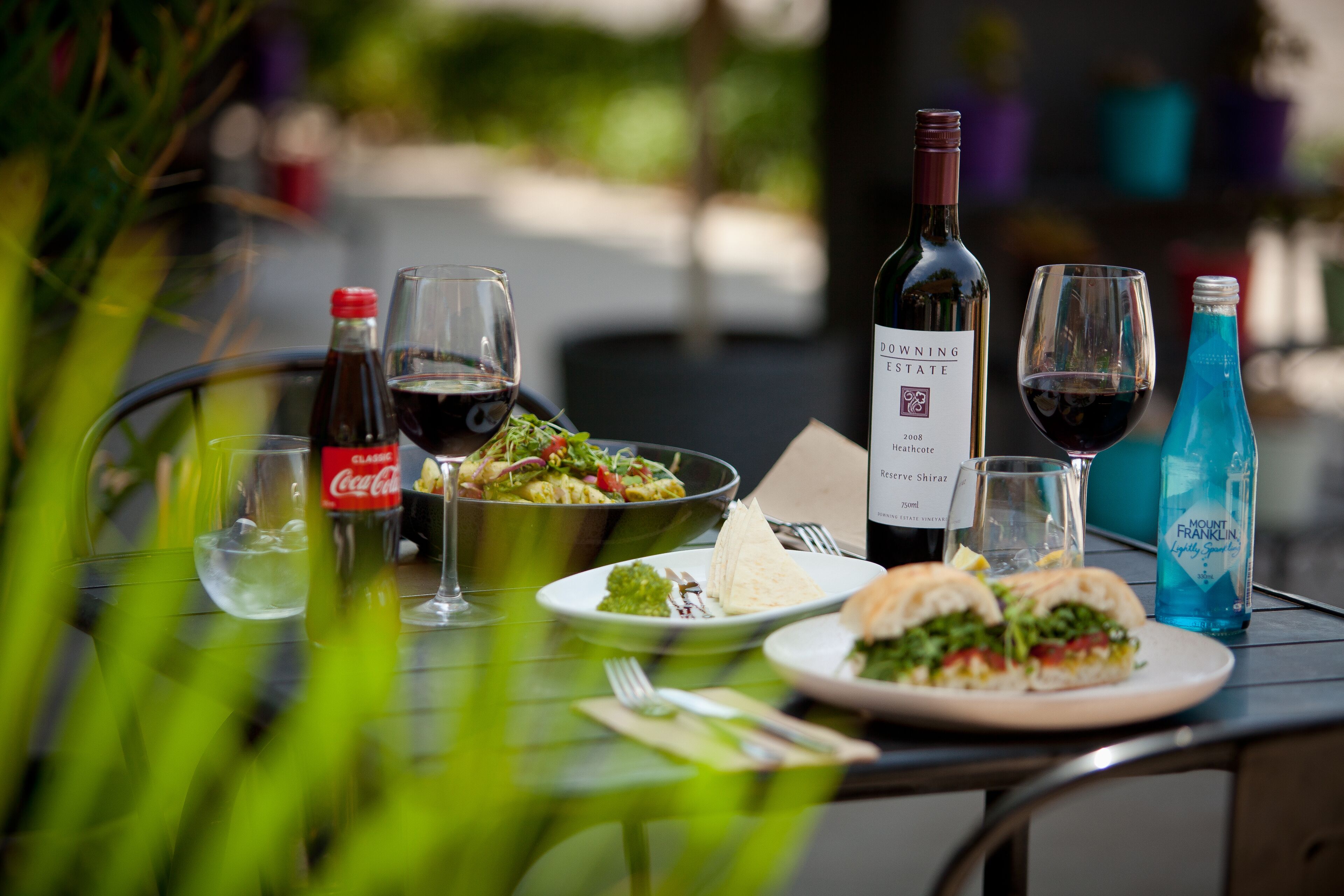 A dining table with a bottle of wine, glasses, a Coca-Cola bottle, a bowl of salad, and a sandwich.