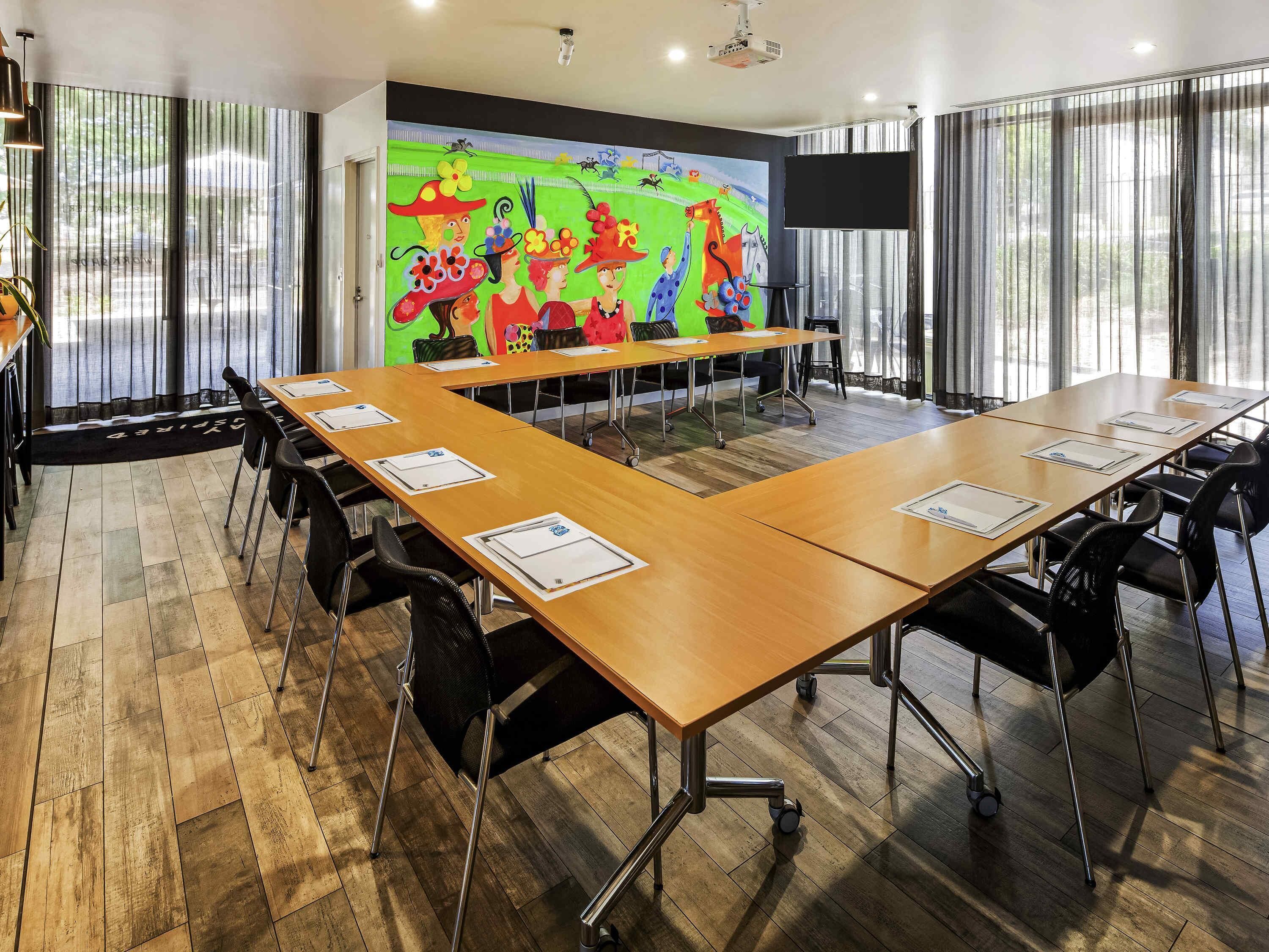 A conference room with a long table, chairs, a projector screen, and a colorful mural on the wall.