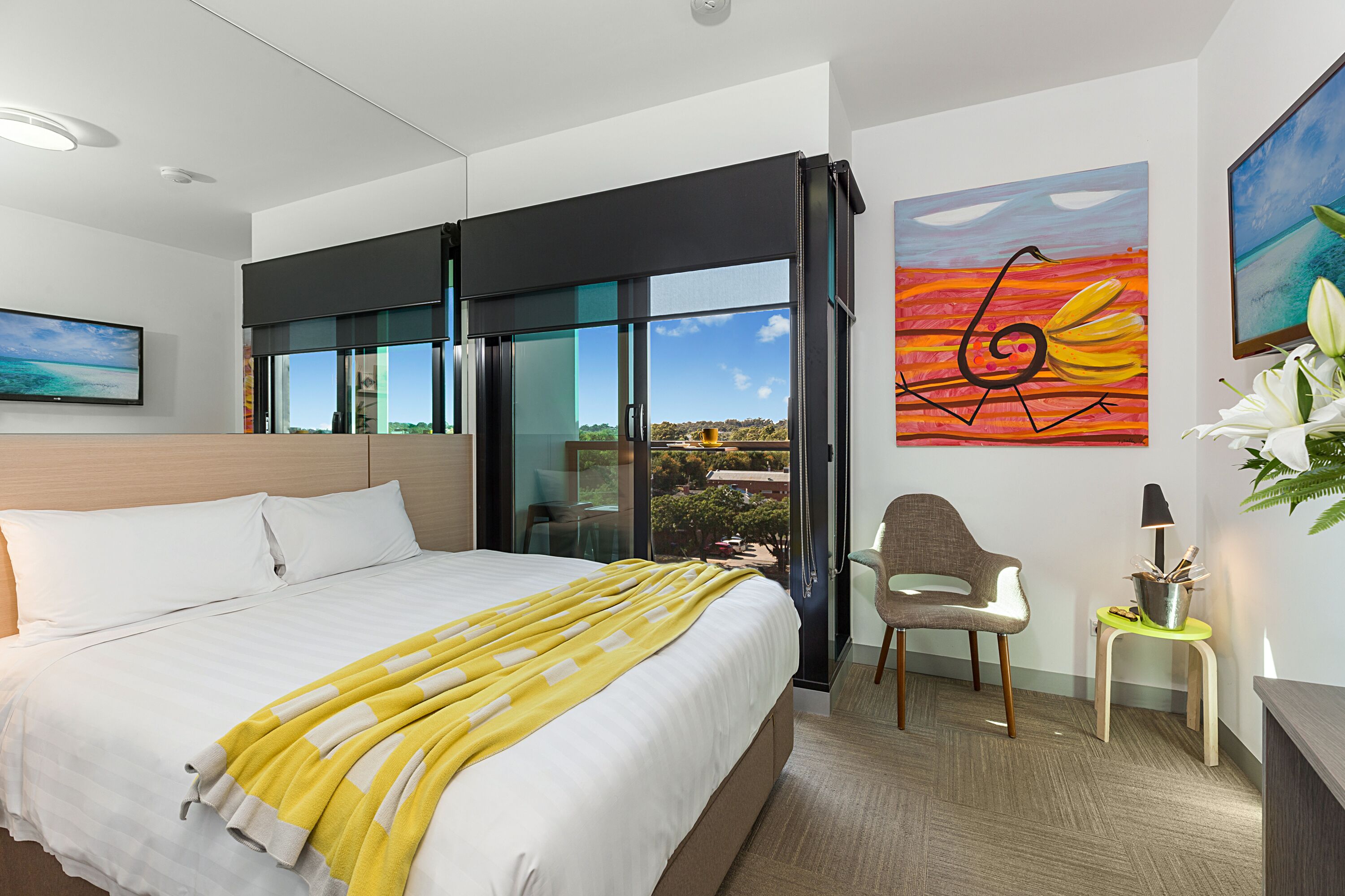 Habitación de hotel moderna con una cama grande, vista a través de una puerta corrediza, una silla, una mesita de noche con una lámpara y obras de arte en las paredes.