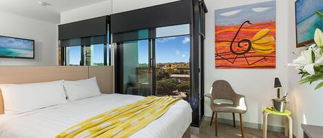 Habitación de hotel moderna con una cama grande, vista a través de una puerta corrediza, una silla, una mesita de noche con una lámpara y obras de arte en las paredes.