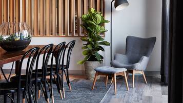 Un comedor moderno con una mesa de madera, sillas negras, una planta en maceta, una lámpara de pie y un sillón gris.