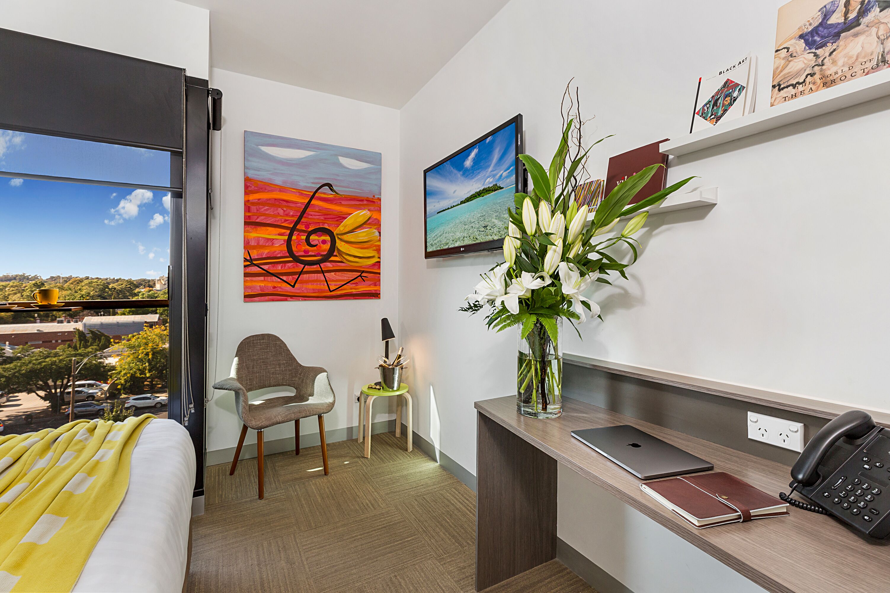 A hotel room with a bed, a desk with a laptop, a phone, and a vase with flowers. There are two paintings on the wall and a window with a view of the city.