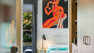 Une chambre d’hôtel avec un lit, un tableau au mur, une fenêtre donnant sur une voiture et des arbres, et une lampe sur une table de chevet.