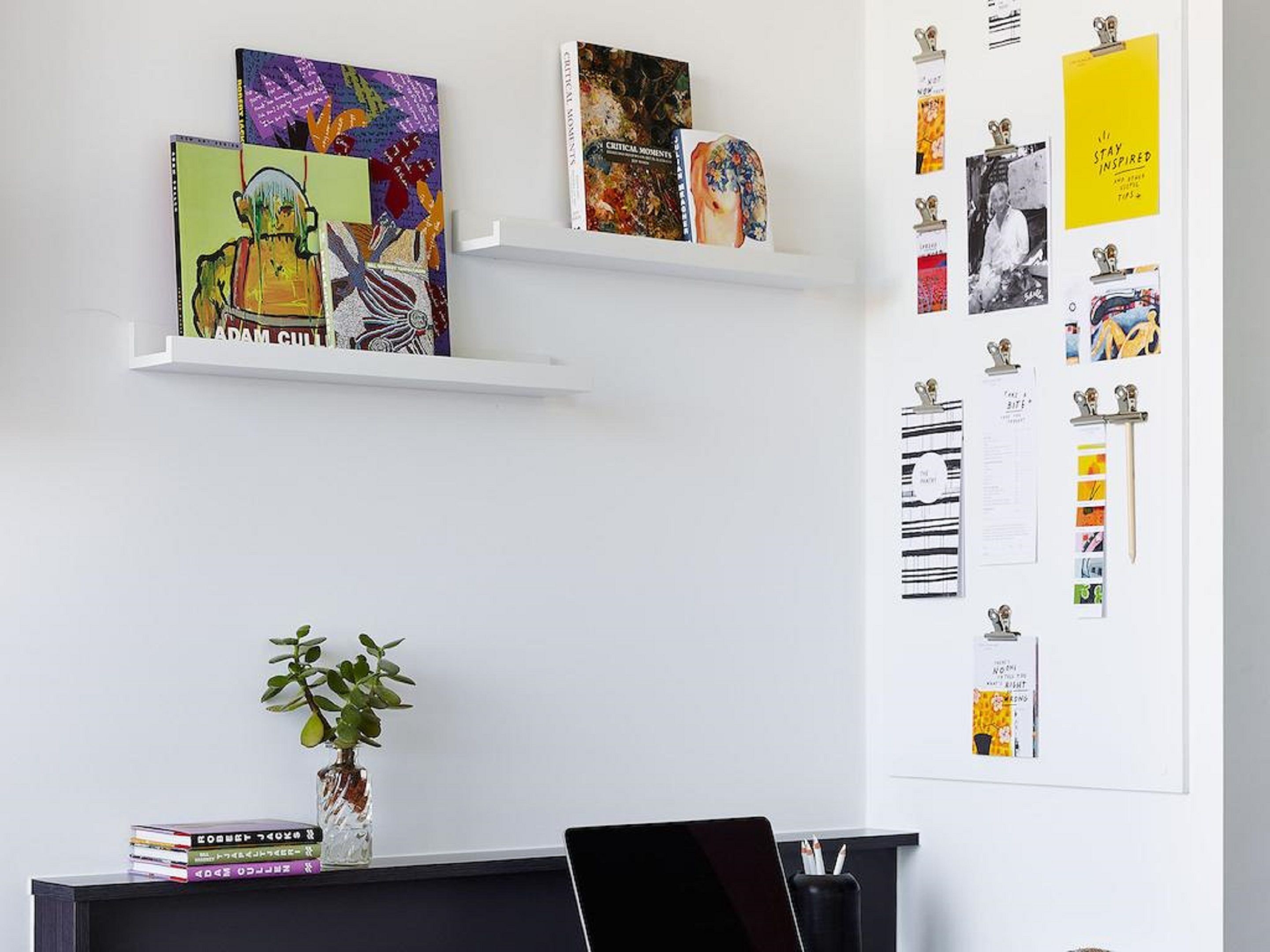 Un espacio de trabajo con dos estanterías blancas que exhiben libros y obras de arte, una planta en maceta, y una pared con papeles y notas sujetas.