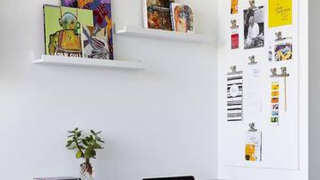 Un espacio de trabajo con dos estanterías blancas que exhiben libros y obras de arte, una planta en maceta, y una pared con papeles y notas sujetas.