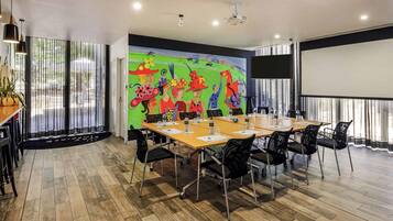 A conference room with a large table, black chairs, a projector screen, and a colorful mural on the wall.