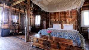 A rustic wooden room with a large bed, a wooden chest, a small table with a lamp, and a window with curtains. - Sala Lodges (Siem Reap)