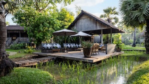 A wooden deck with outdoor furniture and umbrellas, surrounded by lush greenery and a water feature. - Sala Lodges (Siem Reap)