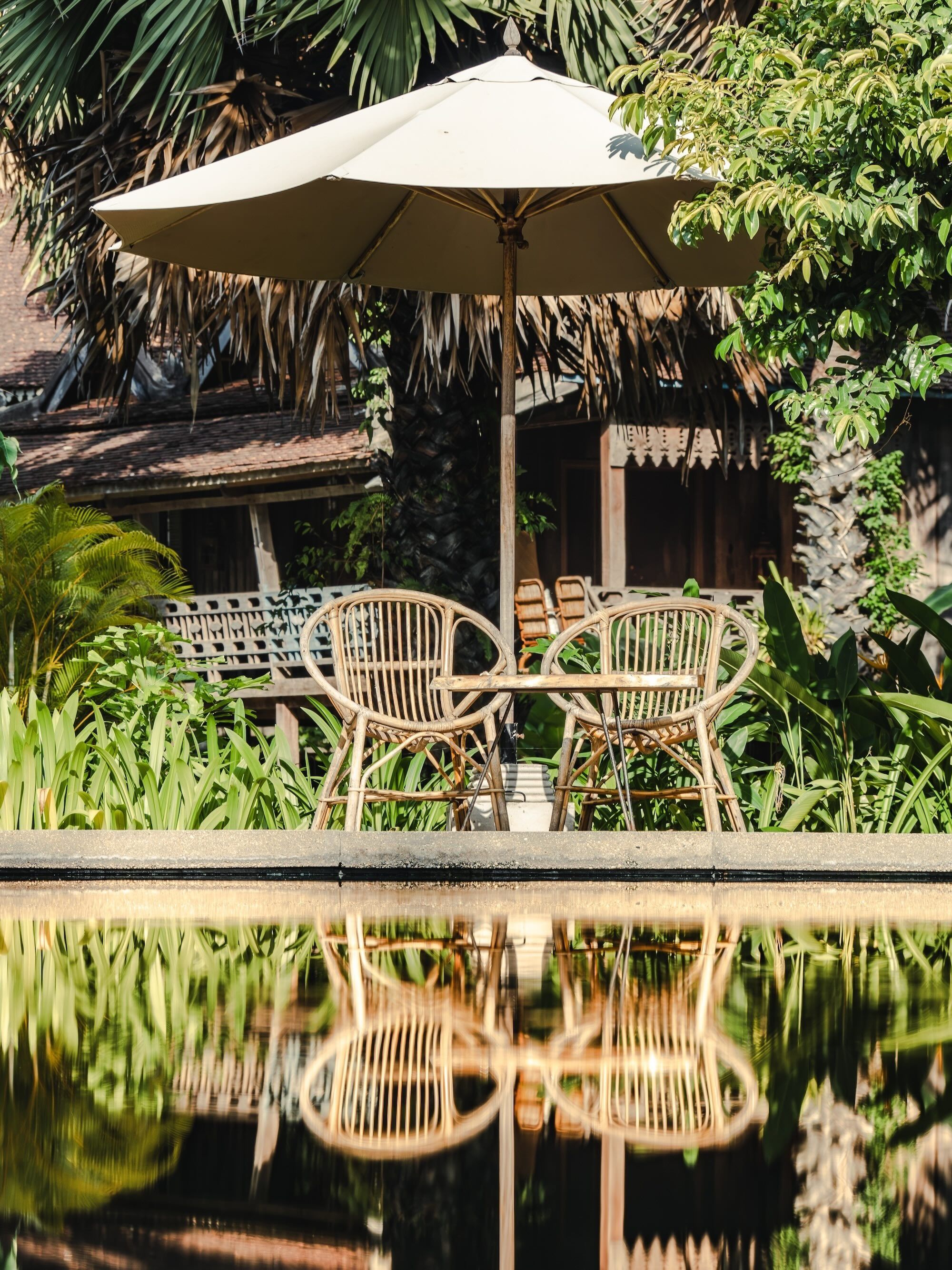 A poolside area with two wicker chairs and a large umbrella, surrounded by lush greenery and a reflection in the water.