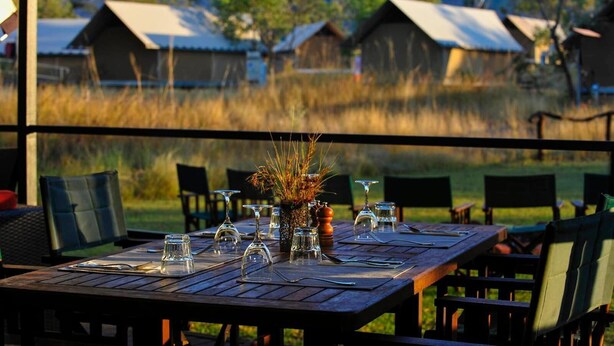 A dining table set outdoors with chairs, a vase with dried plants, and a view of rustic buildings in the background.