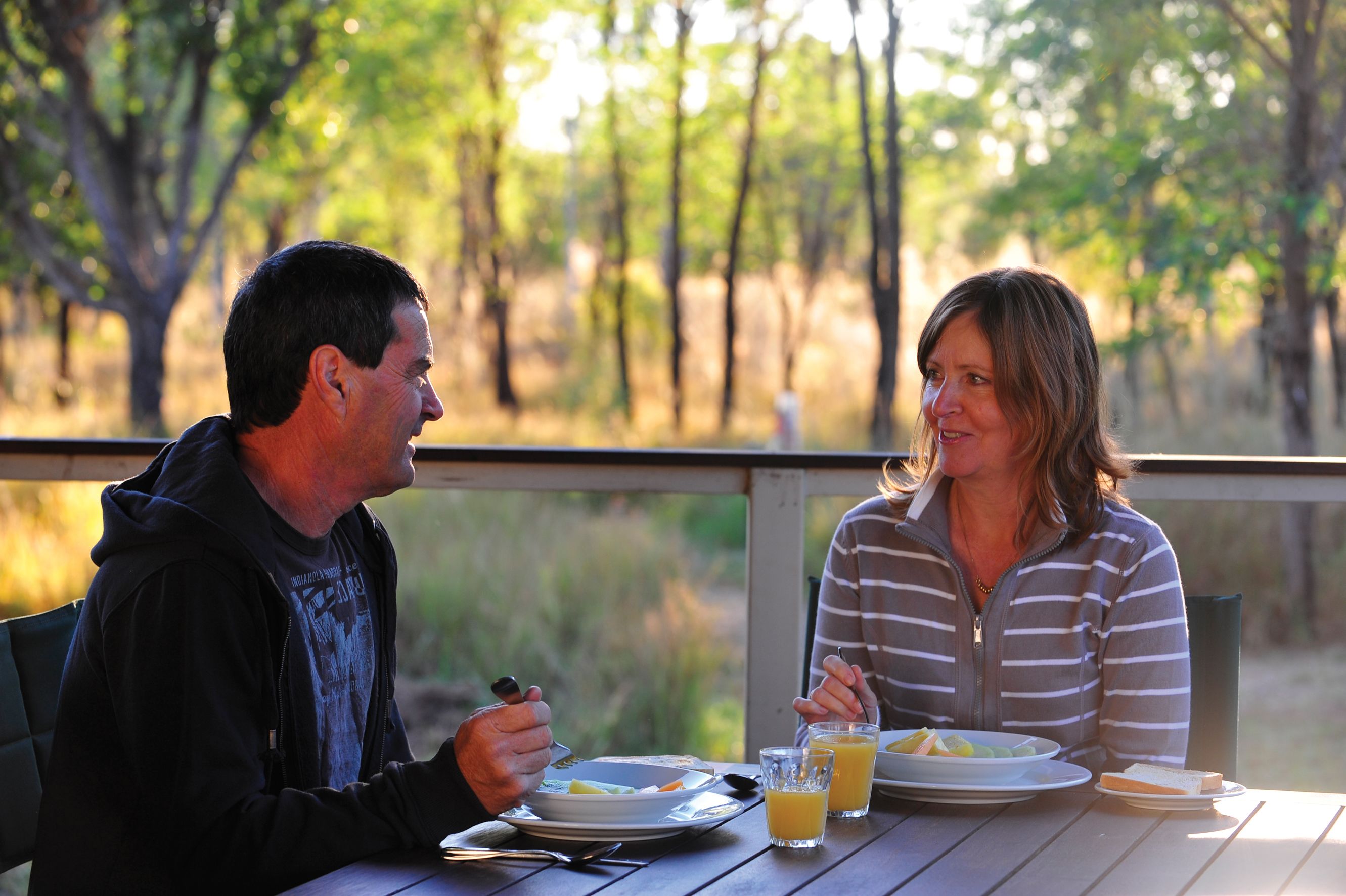 Un couple est assis à une table en plein air, devant des assiettes remplies de nourriture et des boissons, et est entouré d’arbres.