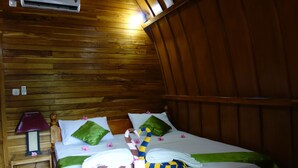 A wooden-paneled room with a bed, pillows, a lamp, and a towel rack. - Banana Leaf Bungalows (Gili Trawangan)