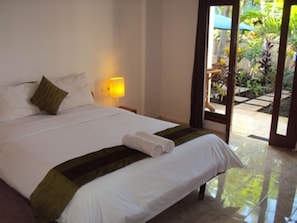 A hotel room with a bed, a nightstand, a lamp, and a view of the outside garden. - Banana Leaf Bungalows (Gili Trawangan)