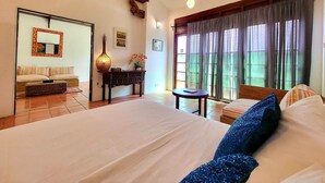 A bedroom with a large bed, a wooden headboard, a bedside table with a lamp, a small table with a book, and a window with curtains. - East Indies Mansion (George Town)