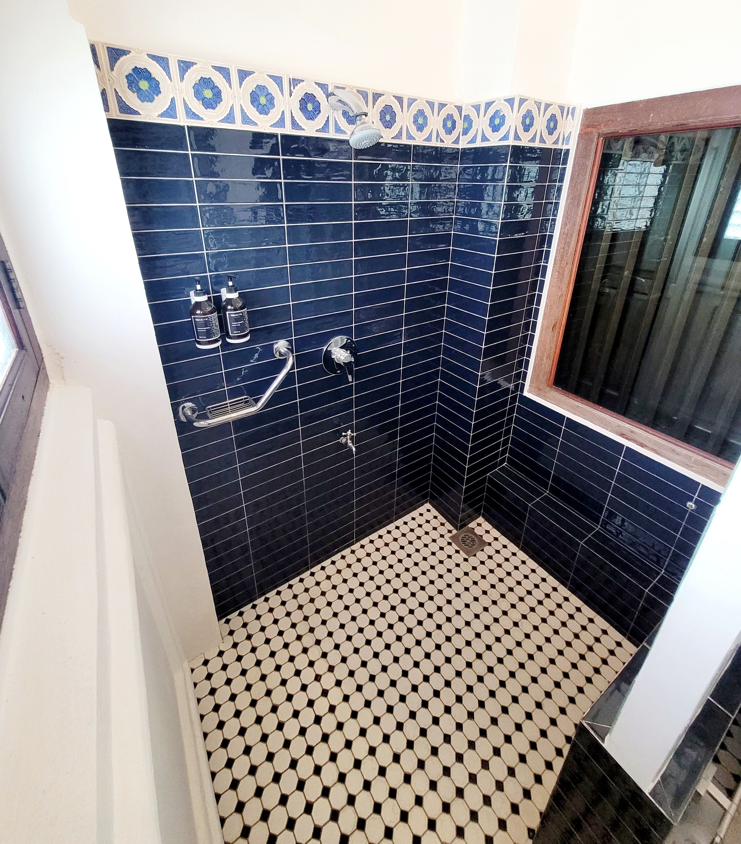 Une salle de bain avec un carrelage noir et blanc à motifs de damier, une douche équipée d’un pommeau de douche à main et une fenêtre avec un cadre en bois.