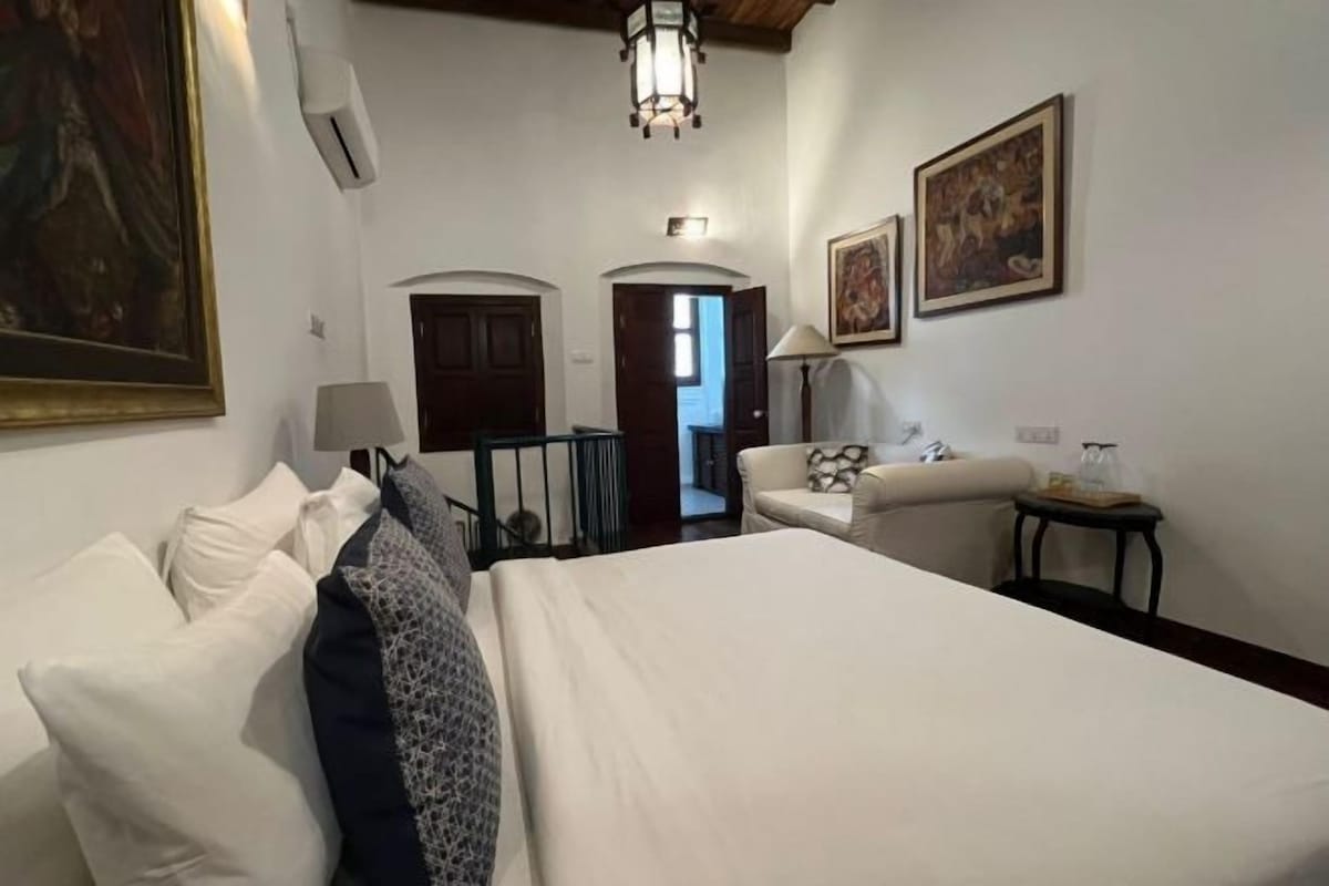 A hotel room with a bed, a sofa, a lamp, and a small table. There are two doors and a hallway leading to another room.