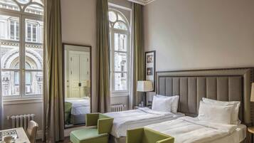 Une chambre d’hôtel avec deux lits, une table à manger et une vue sur un bâtiment historique, visible à travers de grandes fenêtres.