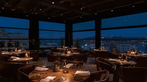 Un restaurant où les tables sont dressées pour le service, de grandes fenêtres offrant une vue sur la ville et un décor intérieur moderne.