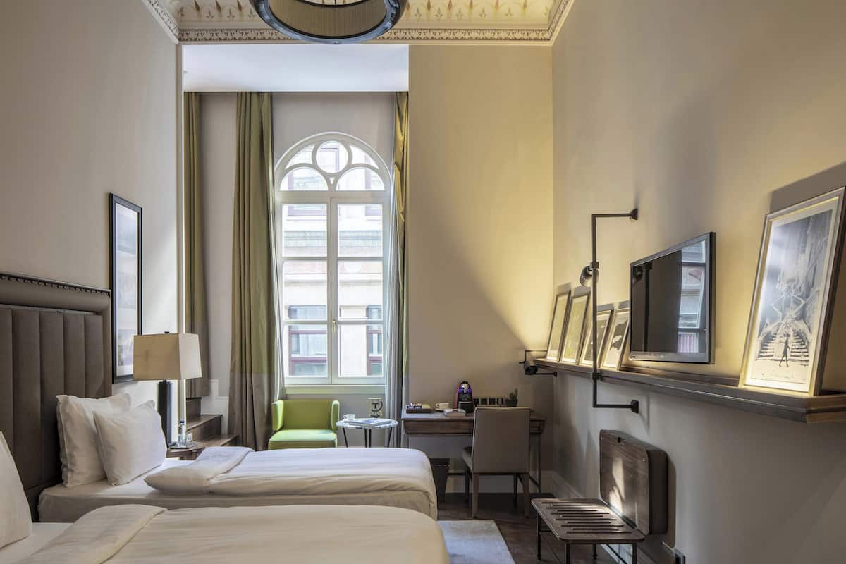 A hotel room with two beds, a desk, a chair, a TV, and a large window with curtains.