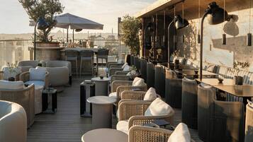A rooftop terrace with wicker furniture, round tables, and hanging lamps.