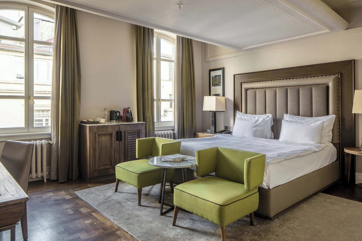 A hotel room with a large bed, two green armchairs, a small round table, a nightstand with a lamp, and a view of the street through large windows.