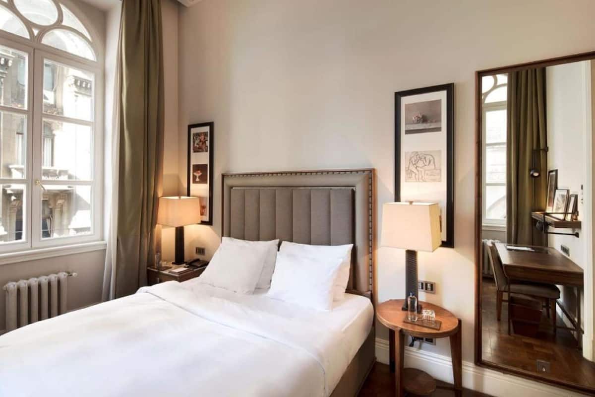 A hotel room with a large bed, a bedside table with a lamp, a small table with a lamp, a mirror, and a view of the street through a window.