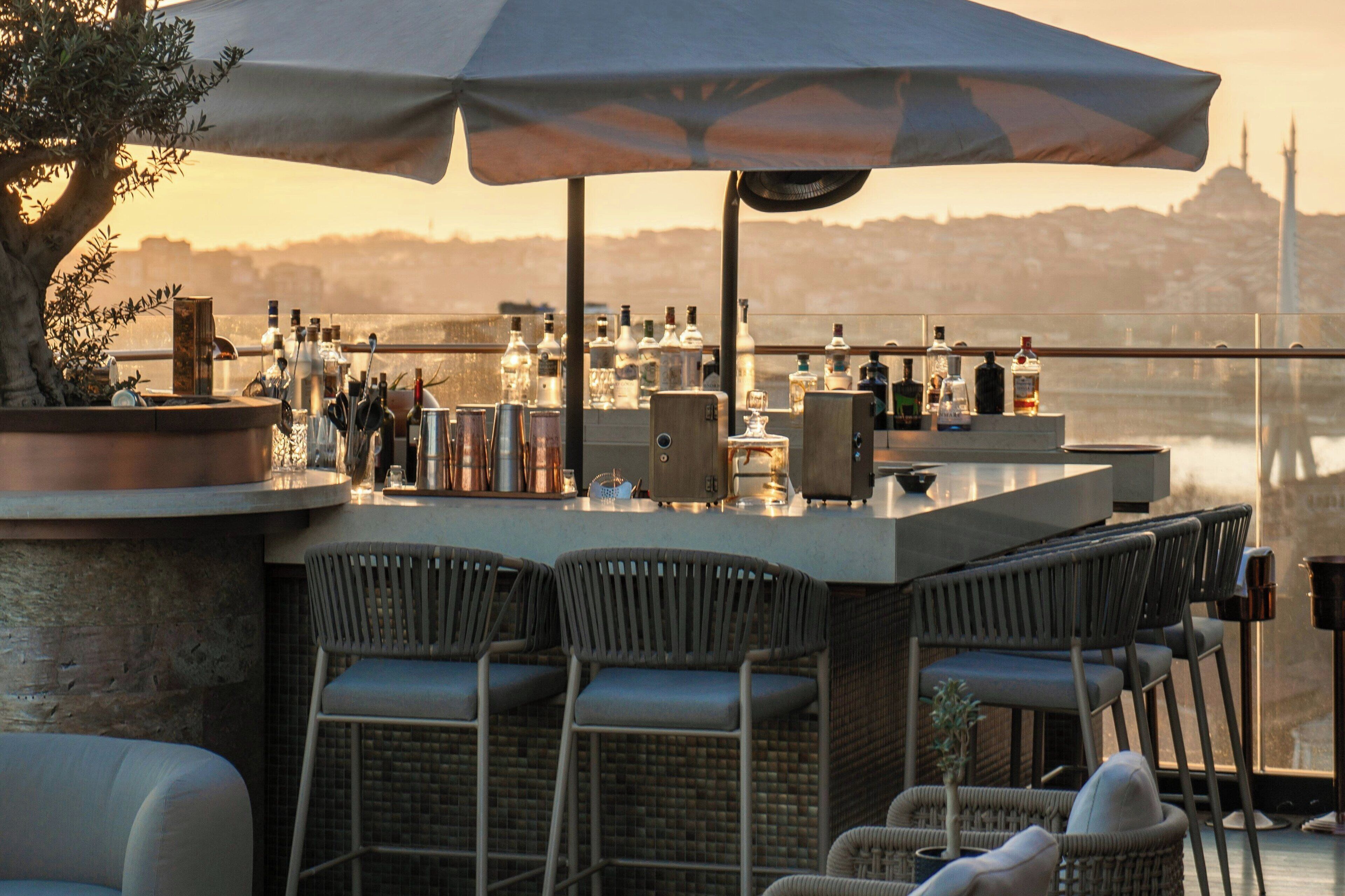 Uma área de bar ao ar livre com cadeiras de vime, um guarda-sol grande e vista para o horizonte da cidade.