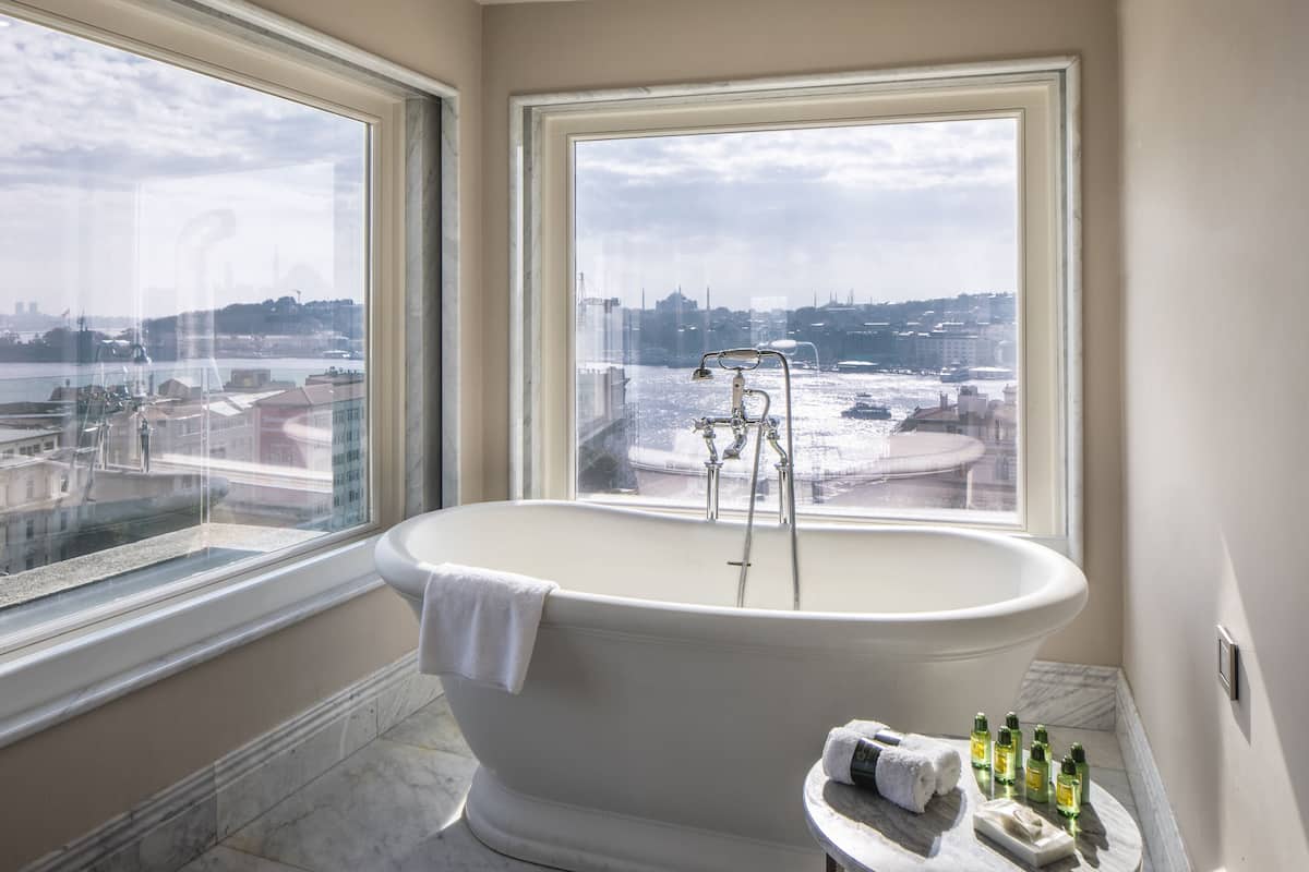 A bathroom with a freestanding bathtub, a window with a city view, and toiletries on a marble countertop.