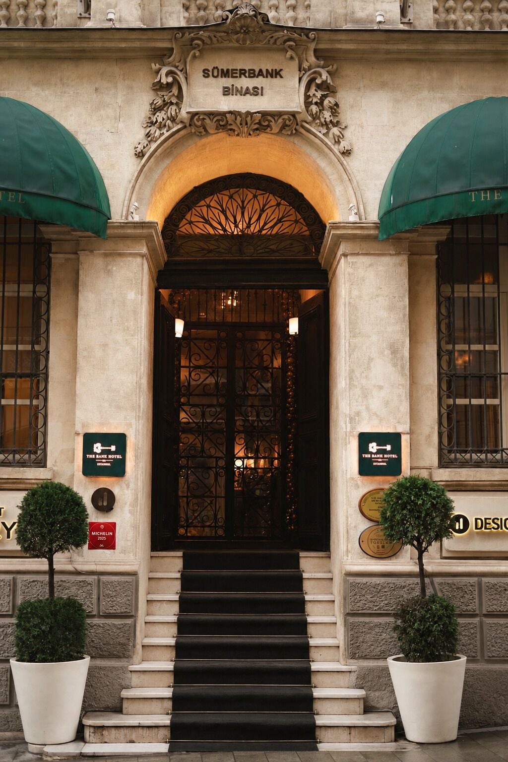 Entrada de un edificio con un letrero que dice 'SÜMERBANK BİNASI' sobre la puerta, una escalera que sube y dos macetas a cada lado.