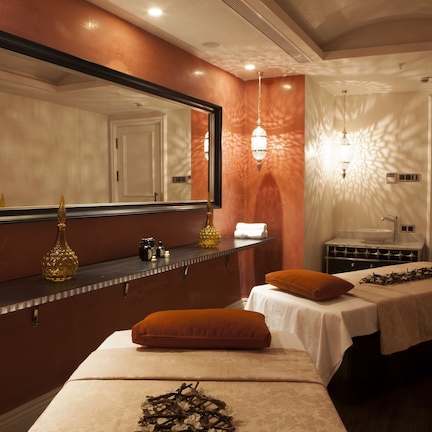 A hotel room with a large mirror, a vanity with toiletries, two beds with white linens, and a wall-mounted light fixture.