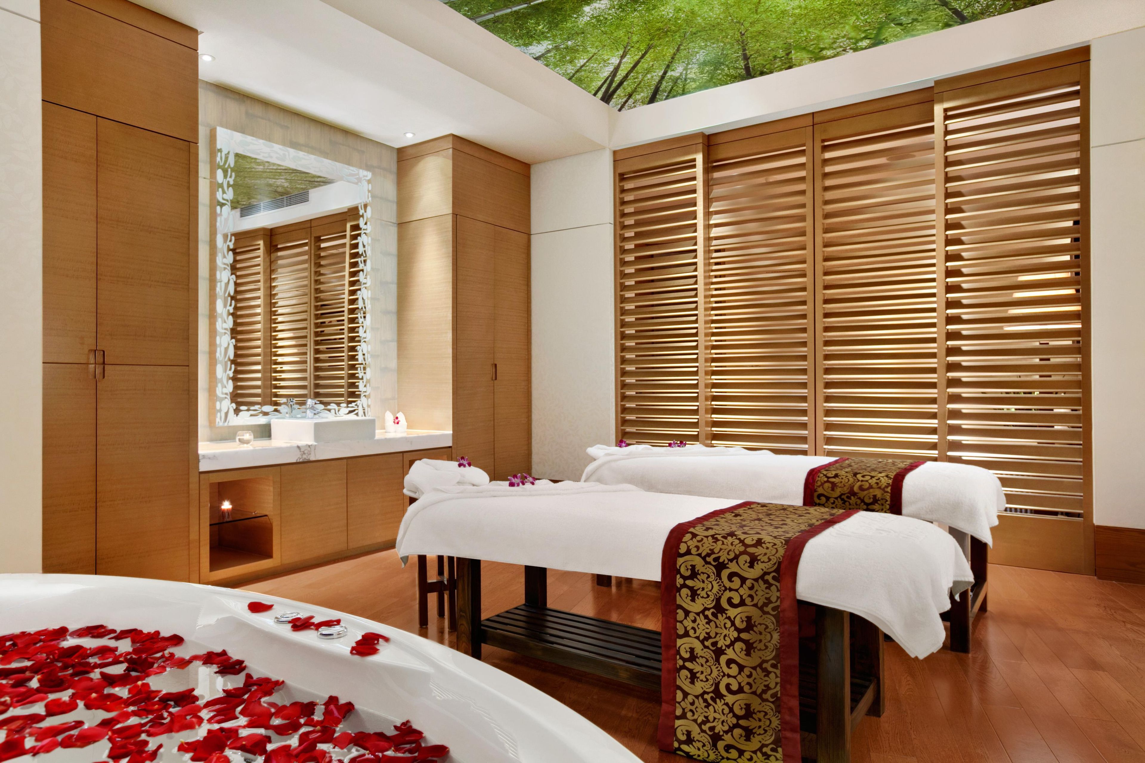 A spa room with two beds, a large bathtub with rose petals, a mirror, and wooden paneling.