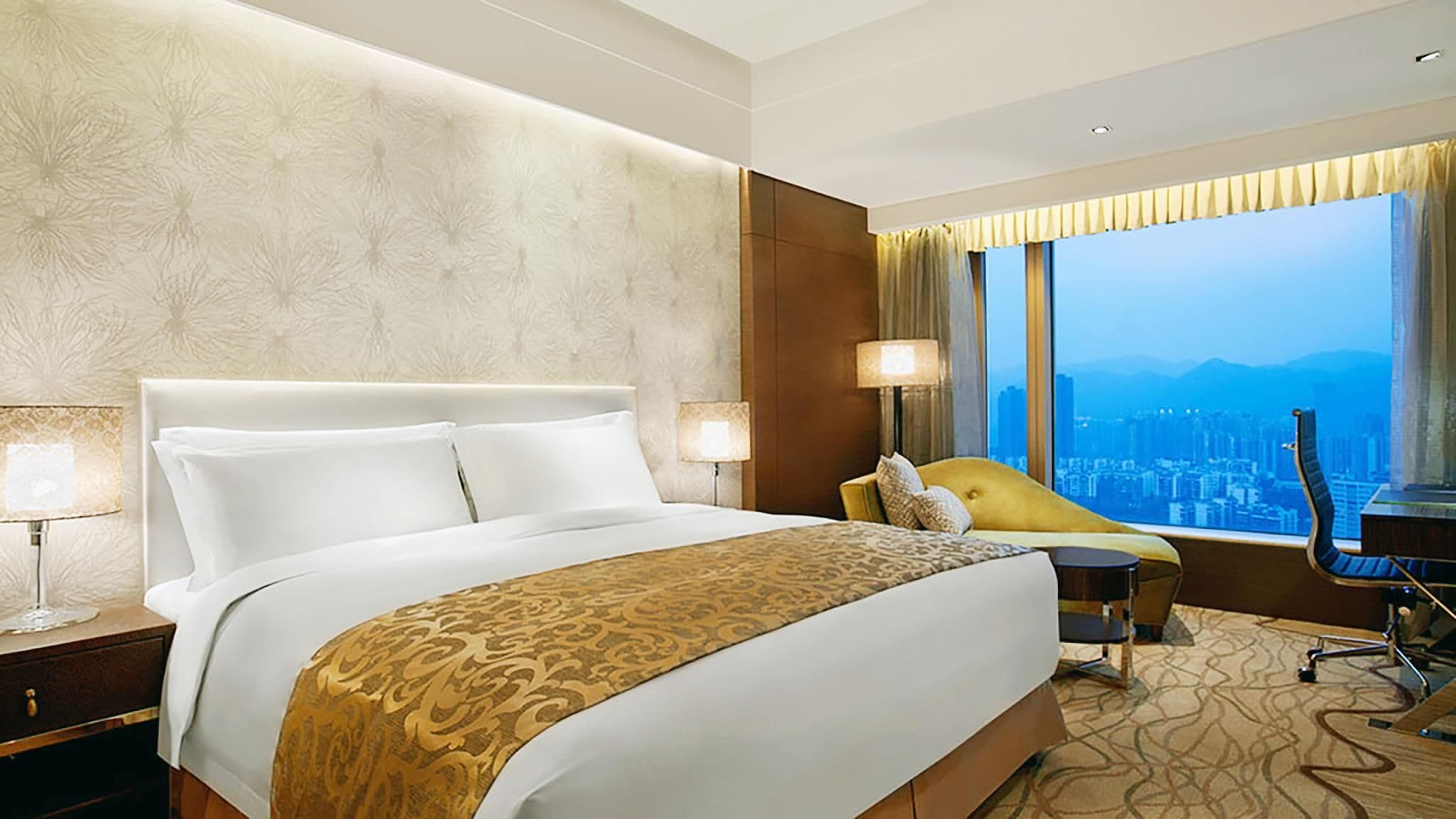 A hotel room with a large bed, a desk, a chair, and a view of a cityscape.