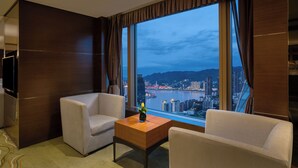 A hotel room with a large window, two white armchairs, a wooden coffee table, and a flat-screen TV.