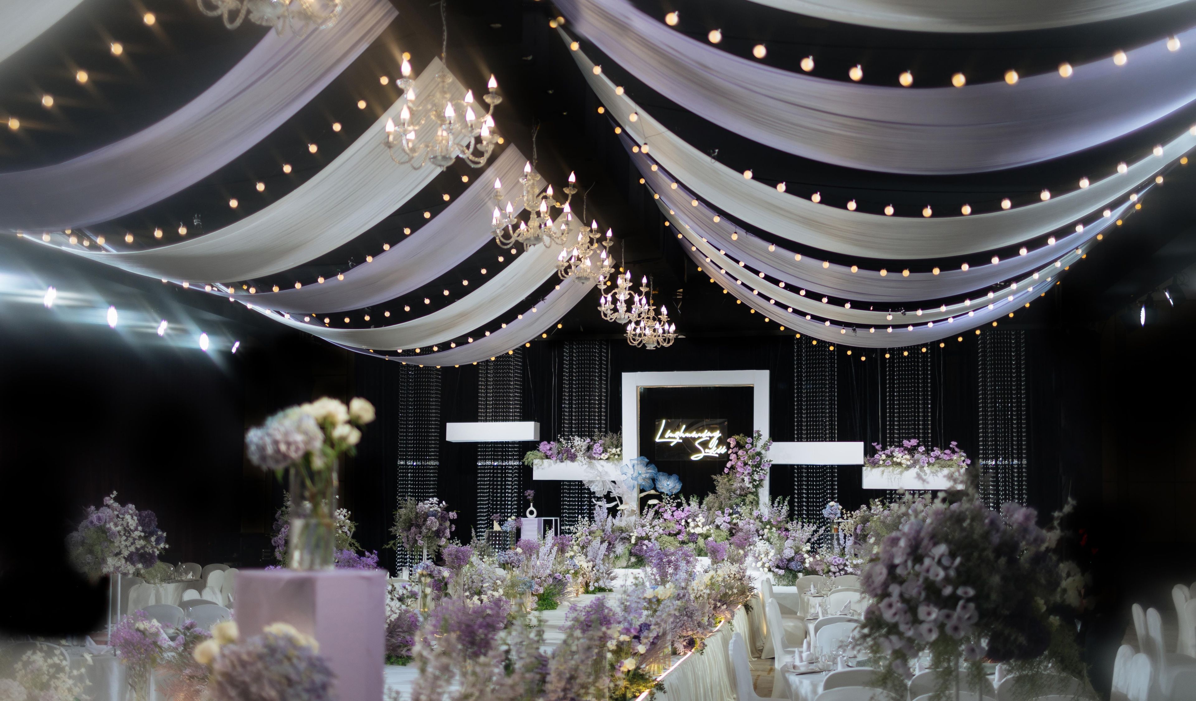 Un salón de banquetes con mesas dispuestas para un evento formal, cubiertas con tela blanca e iluminado por candelabros.