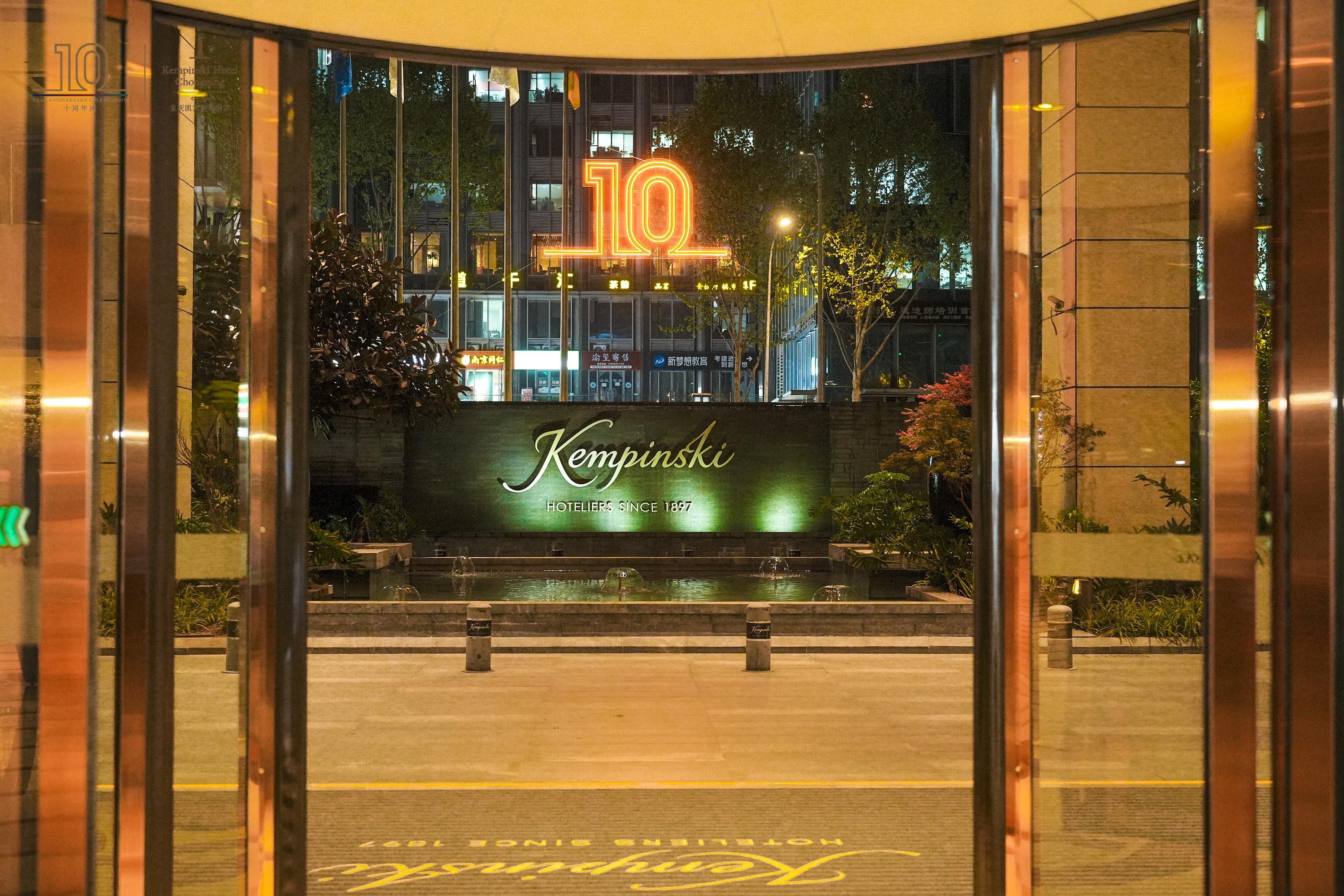 Entrada de un hotel con un gran letrero iluminado con el número '10', una fuente de agua y un cartel que dice 'Kempinski Hotels Desde 1897'.