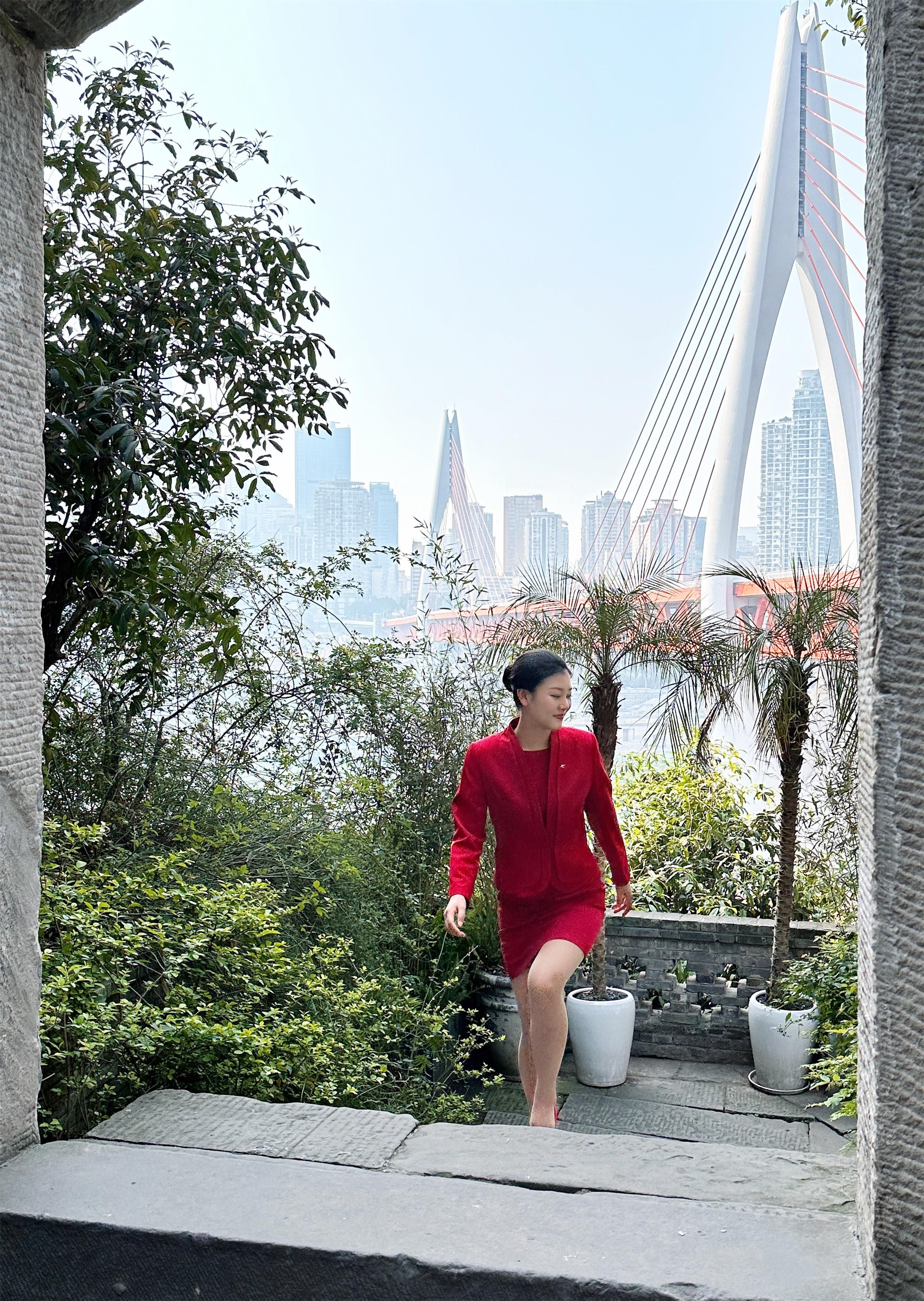 A person in a red dress walking on a stone pathway with a cityscape and a bridge in the background.