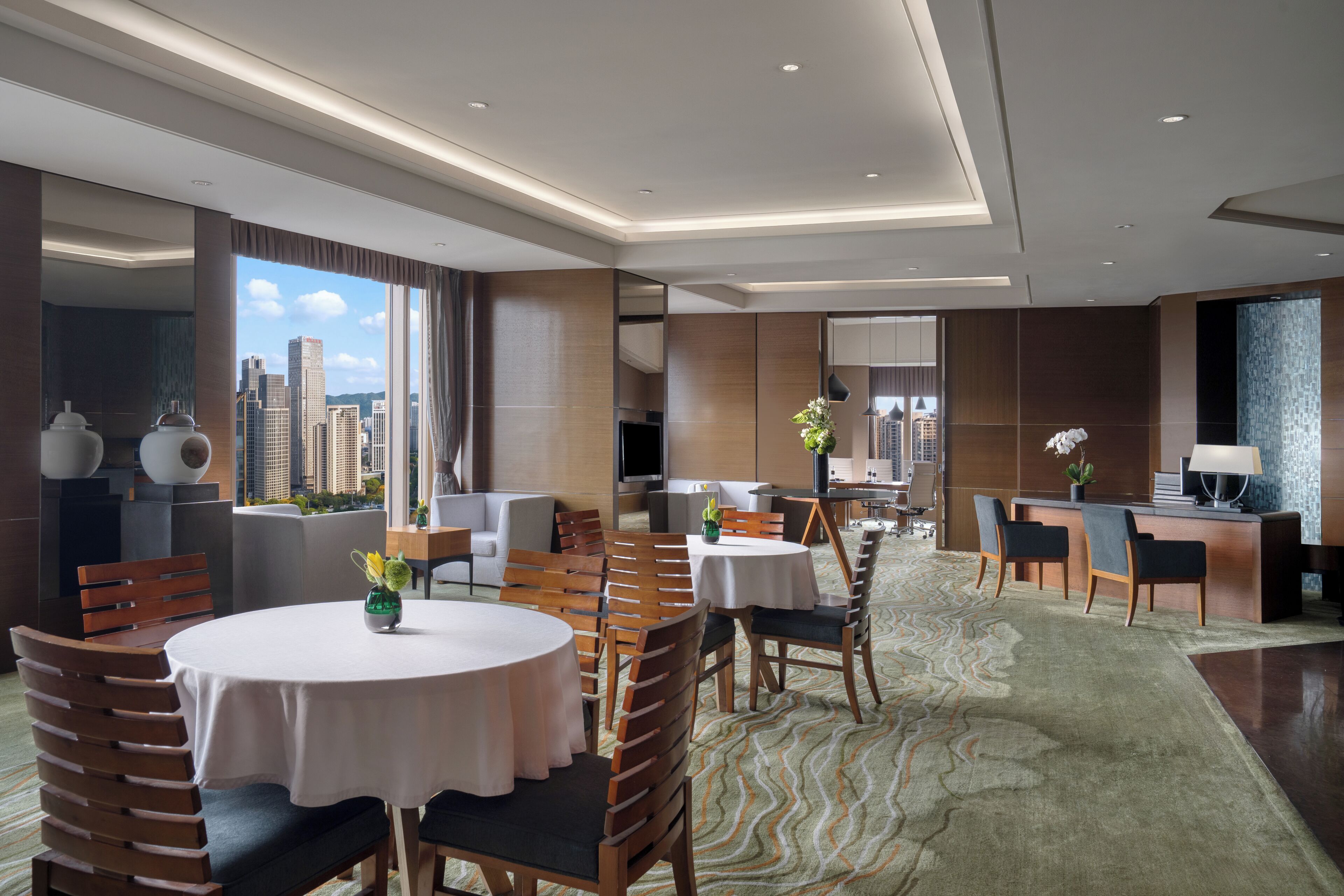 A modern hotel lobby with round tables, wooden chairs, a reception desk, and a city view.